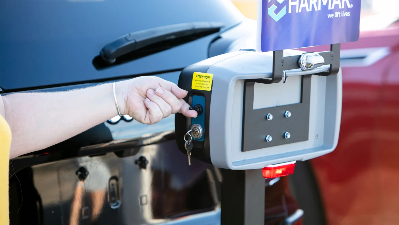 A person operates a key control system on a vehicle lift by Harmar, with a sign above and keys inserted into the lock.